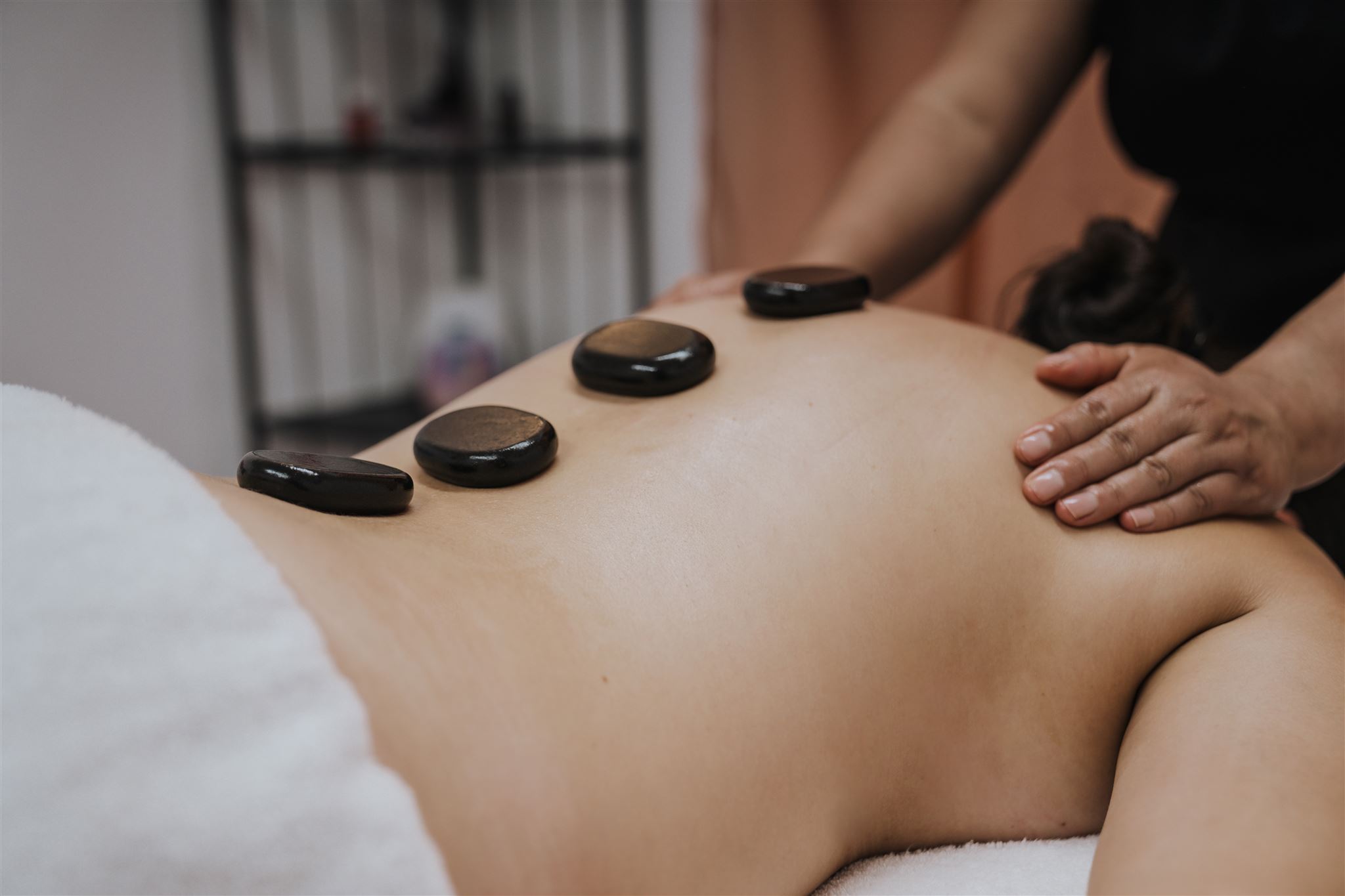 The image depicts a person receiving a massage in a calming setting. Smooth, black stones are placed along their back, likely for a hot stone massage. A therapist's hand is gently applying pressure on the lower back, showcasing a tranquil therapeutic environment.