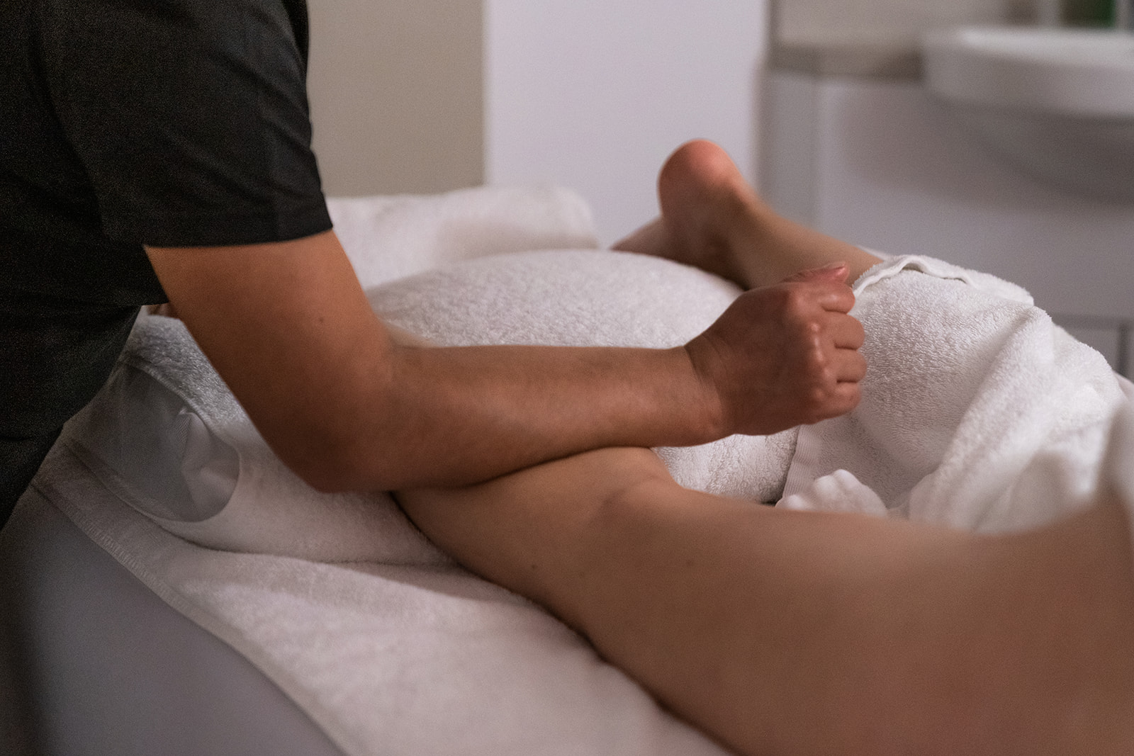 The image showcases a close-up of a massage therapy session. A therapist's hand is gently applying pressure to a client's leg, which is covered with a white towel. The relaxing atmosphere is enhanced by soft lighting, and a comfortable pillow is placed under the leg for support.