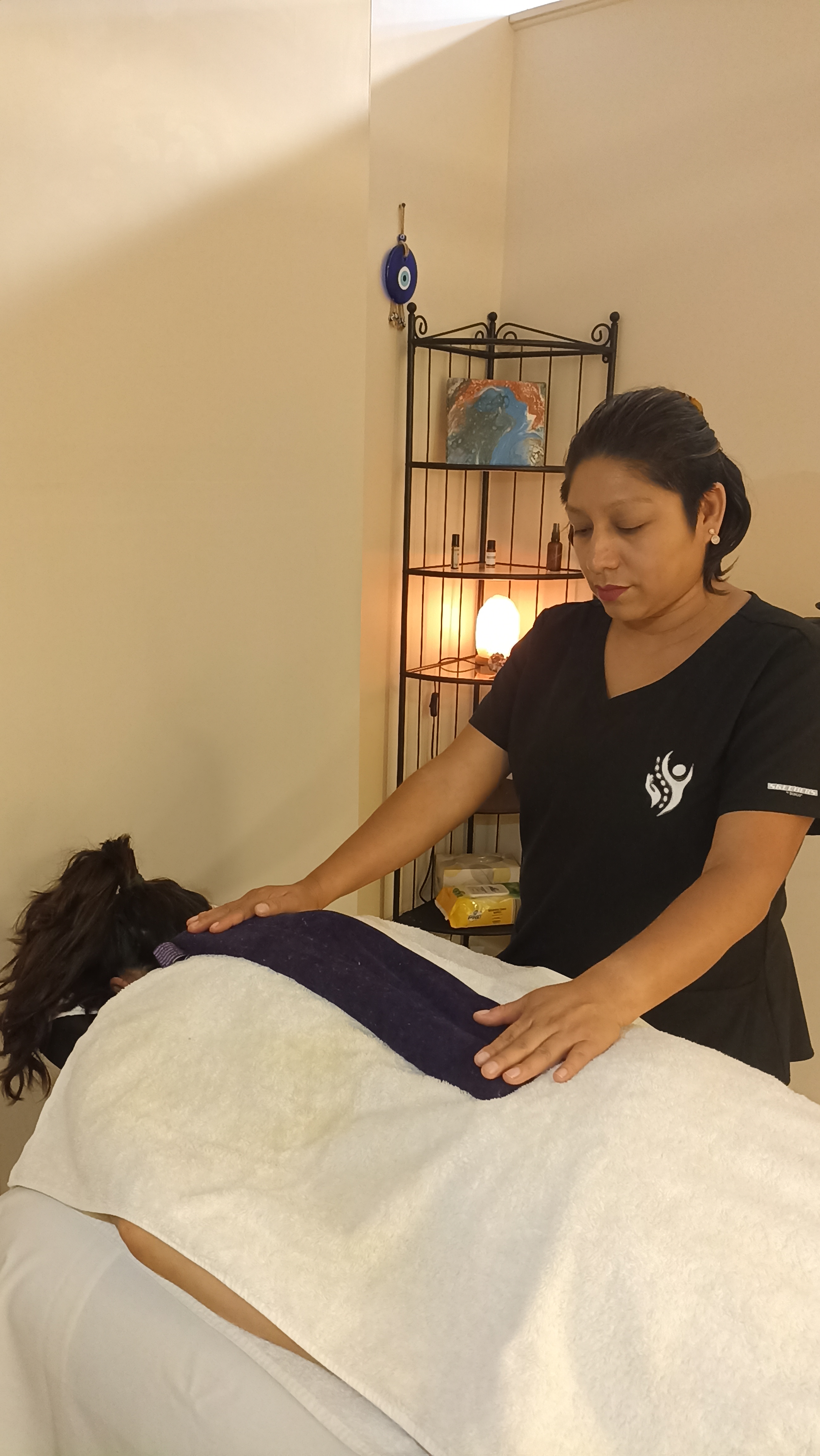 The image shows a massage therapist performing a treatment on a client who is lying face down on a massage table. The therapist is focused and appears to be using a gentle touch, with one hand resting on the client's back and the other preparing to apply a warm towel. The room has a calming ambiance, featuring soft lighting and a decorative stand with a globe and another item, likely a salt lamp, which adds to the relaxing atmosphere.