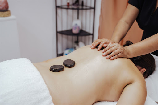 The image shows a person receiving a massage on their back. The therapist is using smooth, heated stones positioned on the person's back, which is covered by a white towel. The scene is calm and relaxing, suggesting a serene spa environment with soft lighting and minimalistic decor in the background.